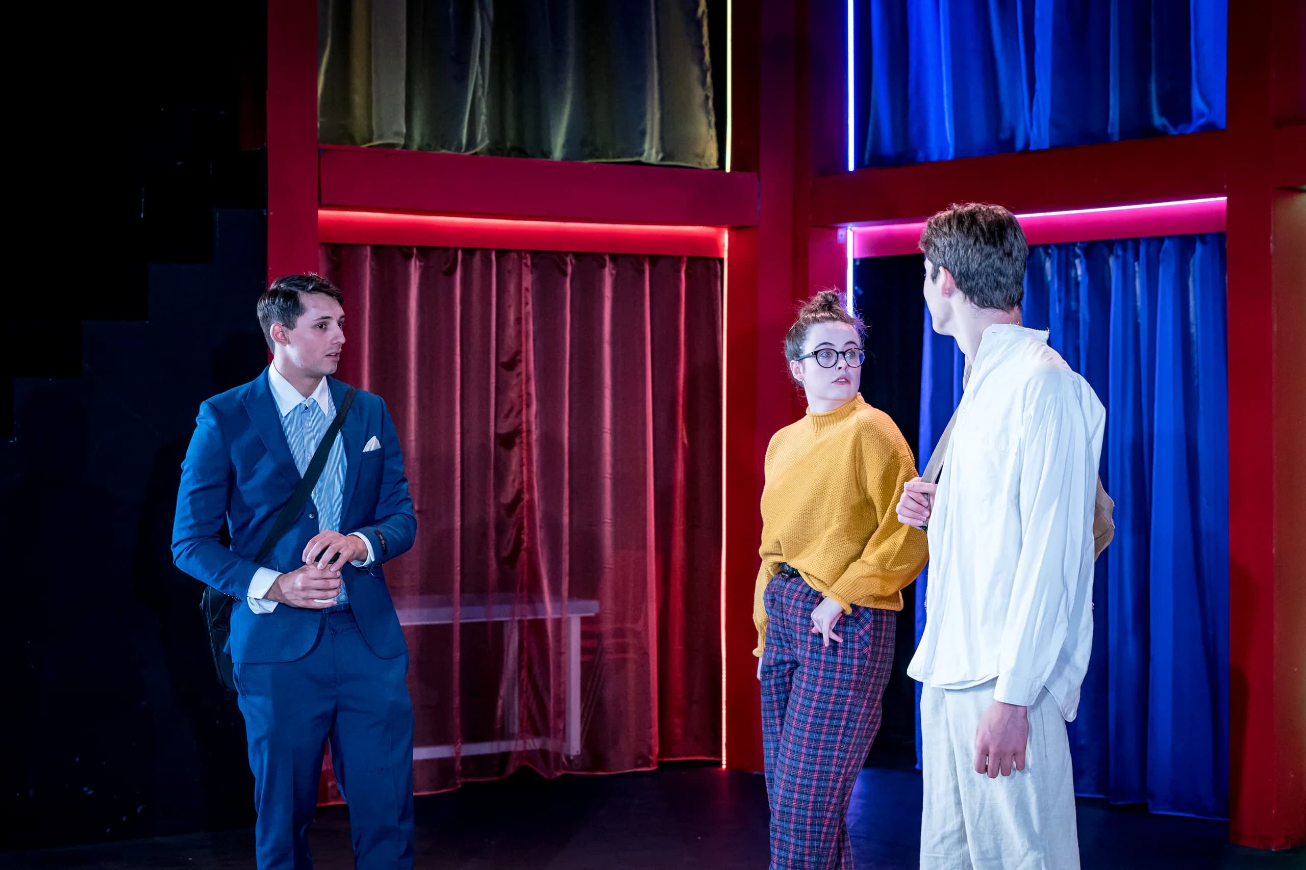 Three actors converse on a neon-lit set of red frames and draped curtains in GSA’s 13.