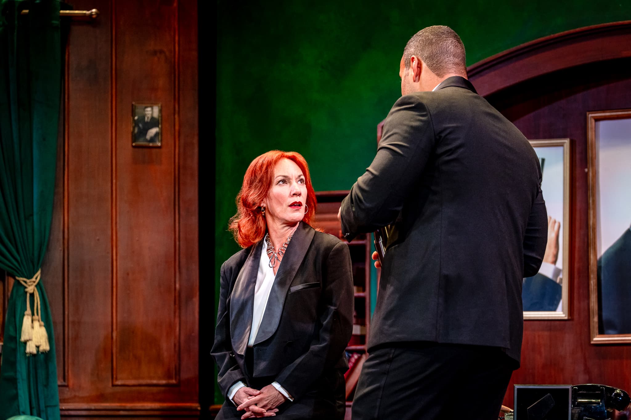 Seated figure in tux-style suit looks up at a standing figure in black; tense exchange in a green, wood‑panelled office set, warm light.