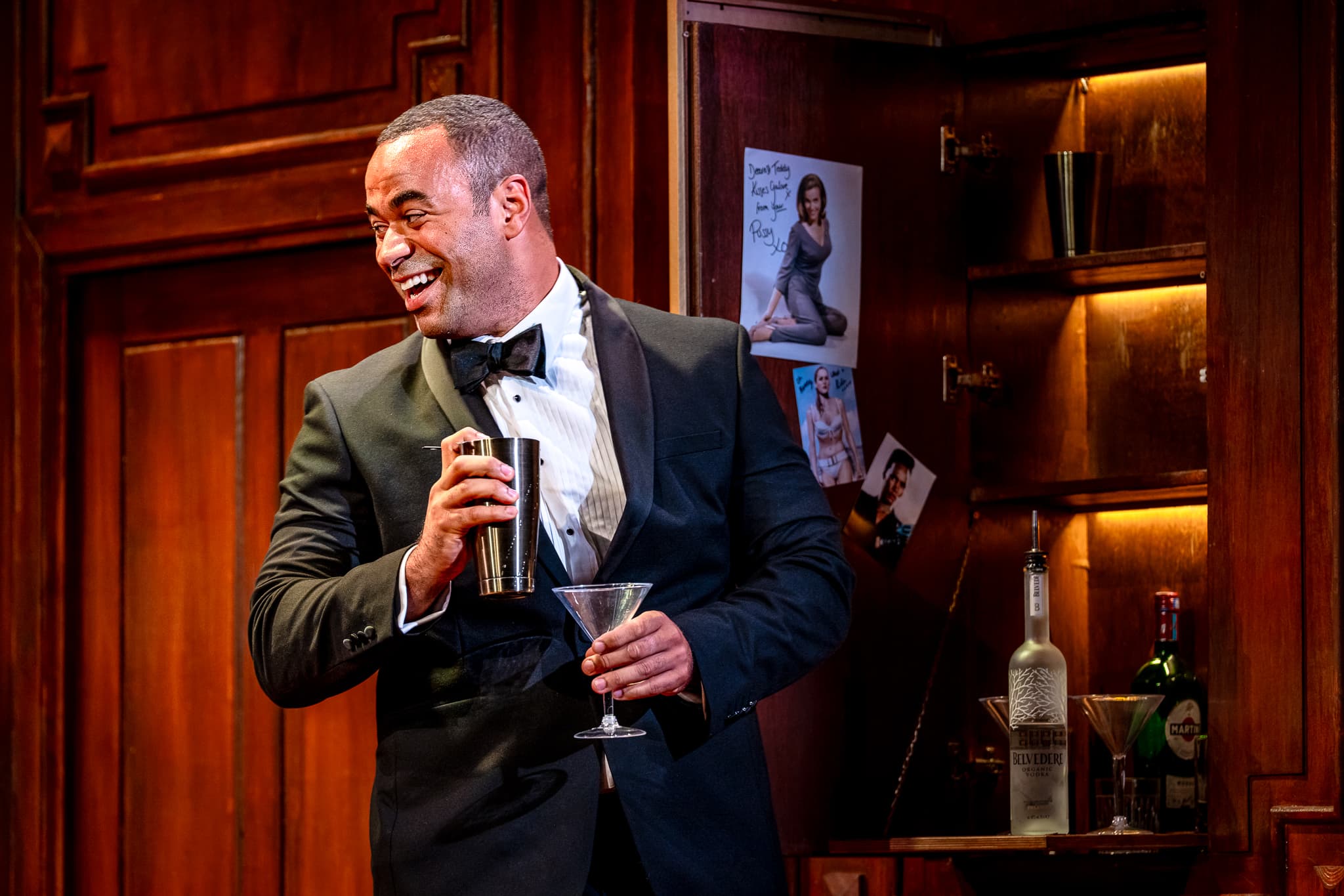 Tuxedoed performer laughs while shaking a martini beside a warm backlit drinks cabinet on a wood‑panelled set.