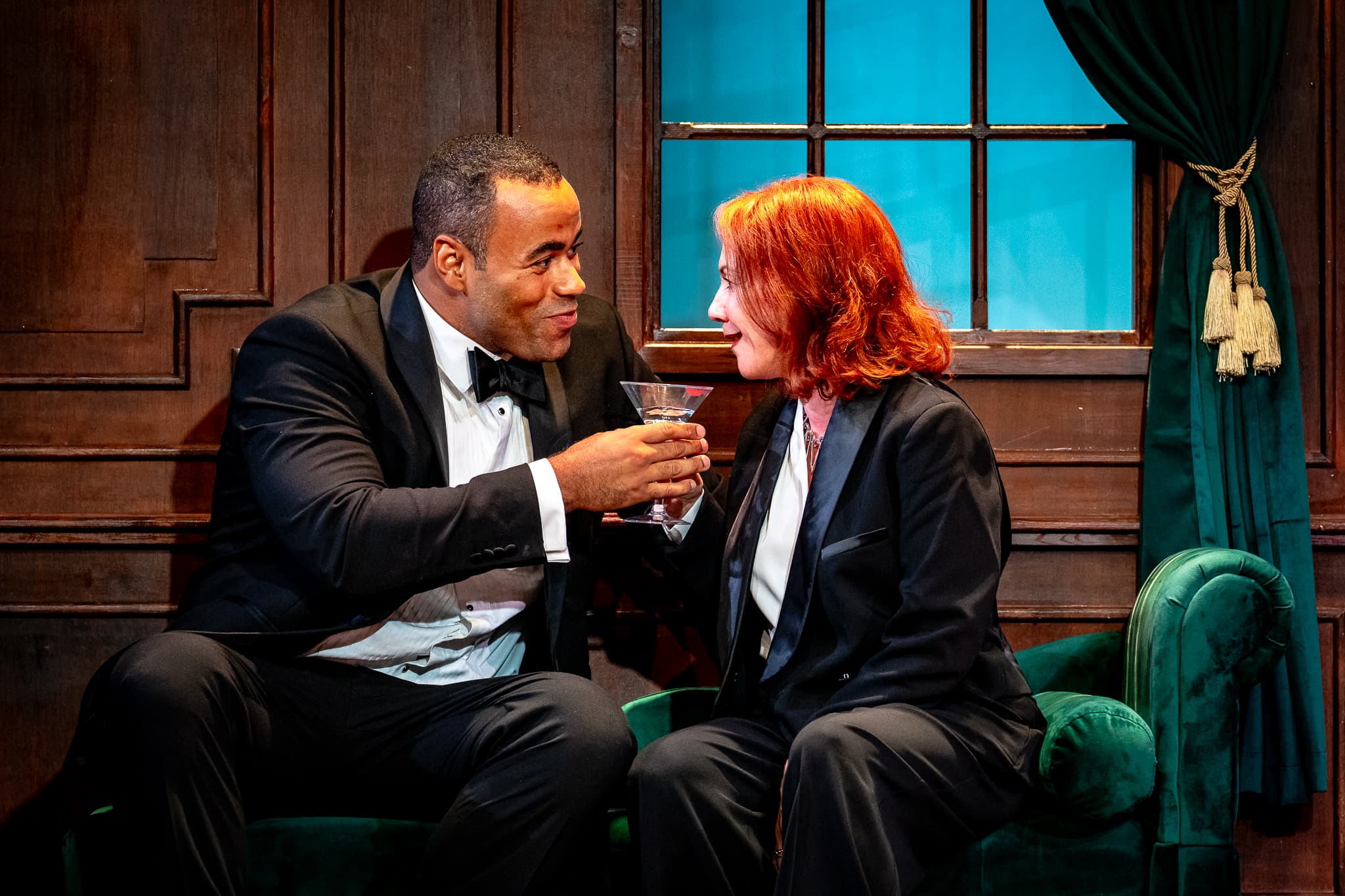 Two tuxedoed characters share a martini on a green velvet sofa; wood‑panelled set with teal‑lit window, playful mood.