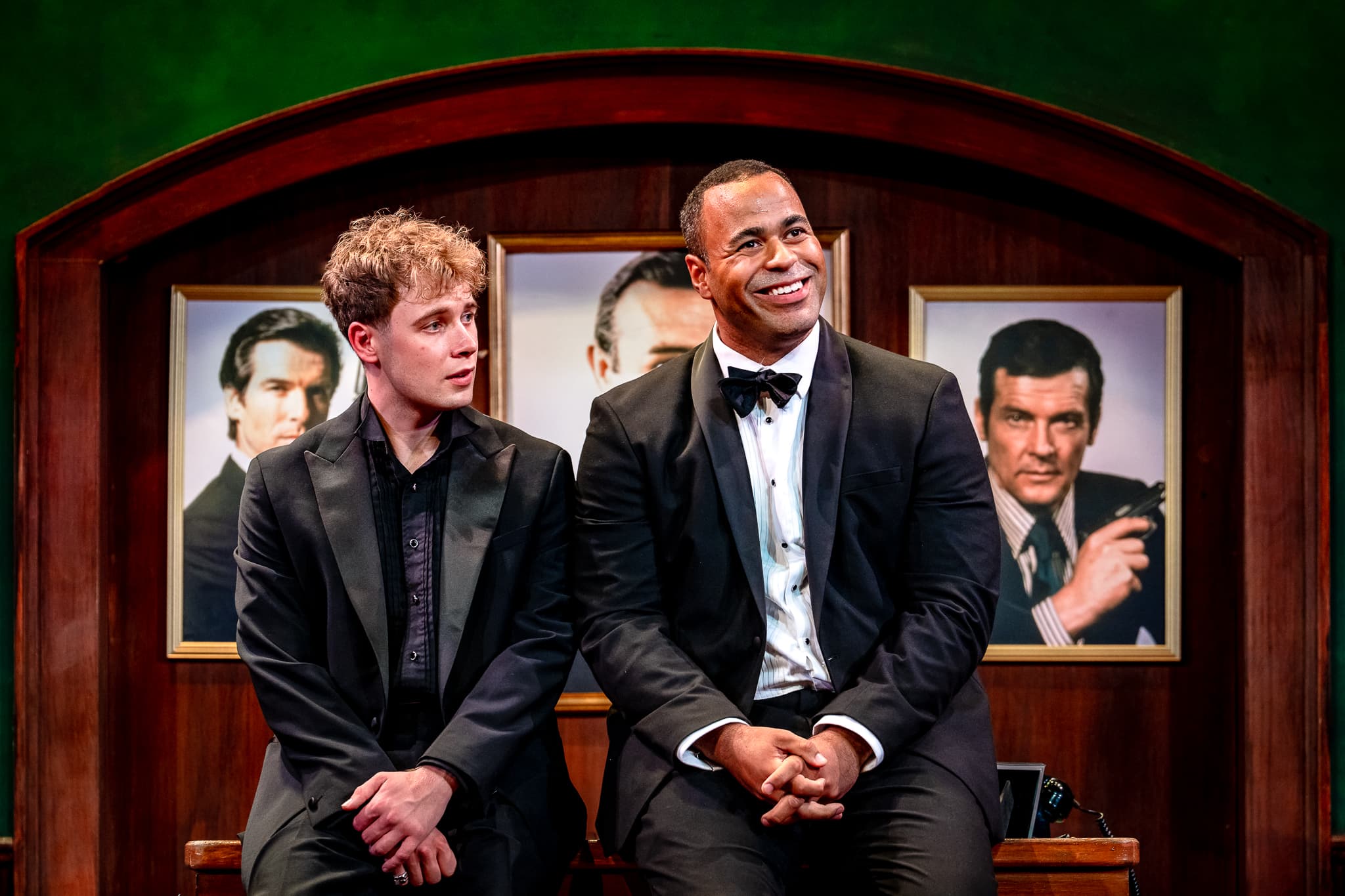 Two performers in tuxedos sit on a wooden set beneath three framed portraits; one smiles while the other watches, in A Role To Die For.
