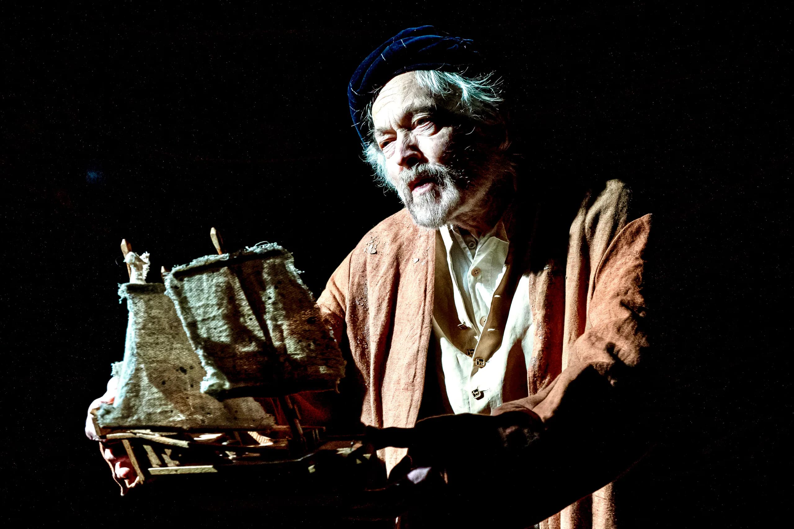 Older performer in robe and cap holds a ragged-sail model ship under stark side light during The Tempest at Jermyn Street Theatre.