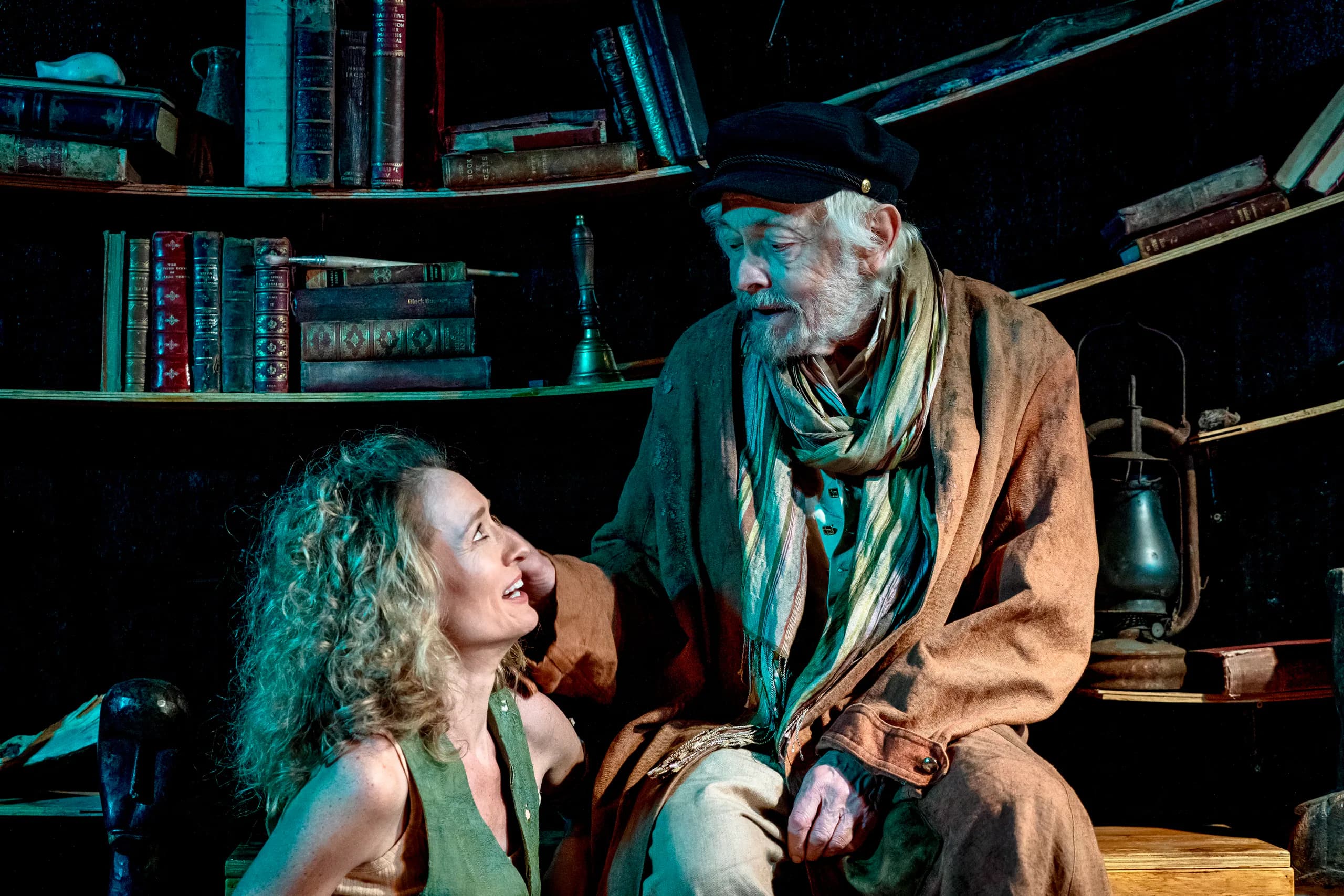 Older man in worn coat and scarf reaches to kneeling woman before curved shelves of books and a lantern; intimate, sea-toned lighting.