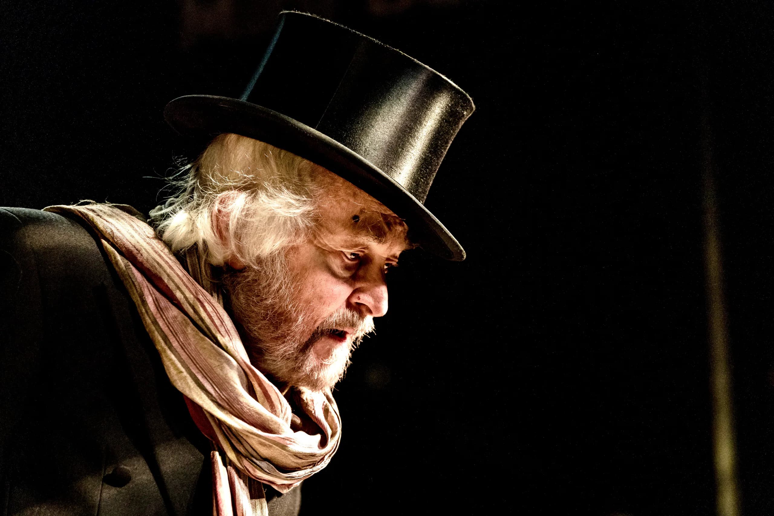 Close-up of an older performer in a top hat and scarf, warm side light picking his profile against a dark stage