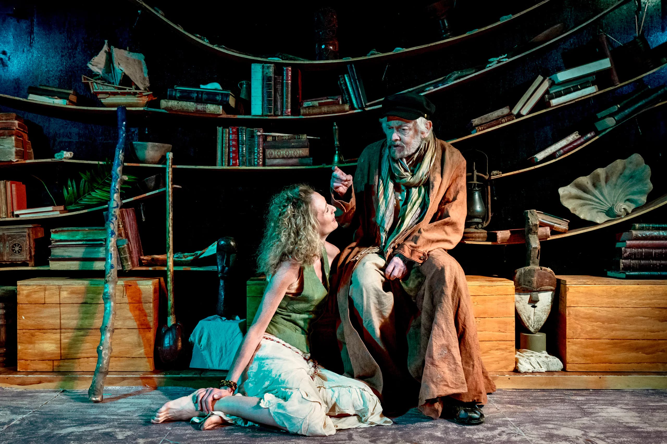 Two performers confer by curved bookshelves; older figure seated, younger kneeling, staff nearby; warm lighting creates an intimate mood.
