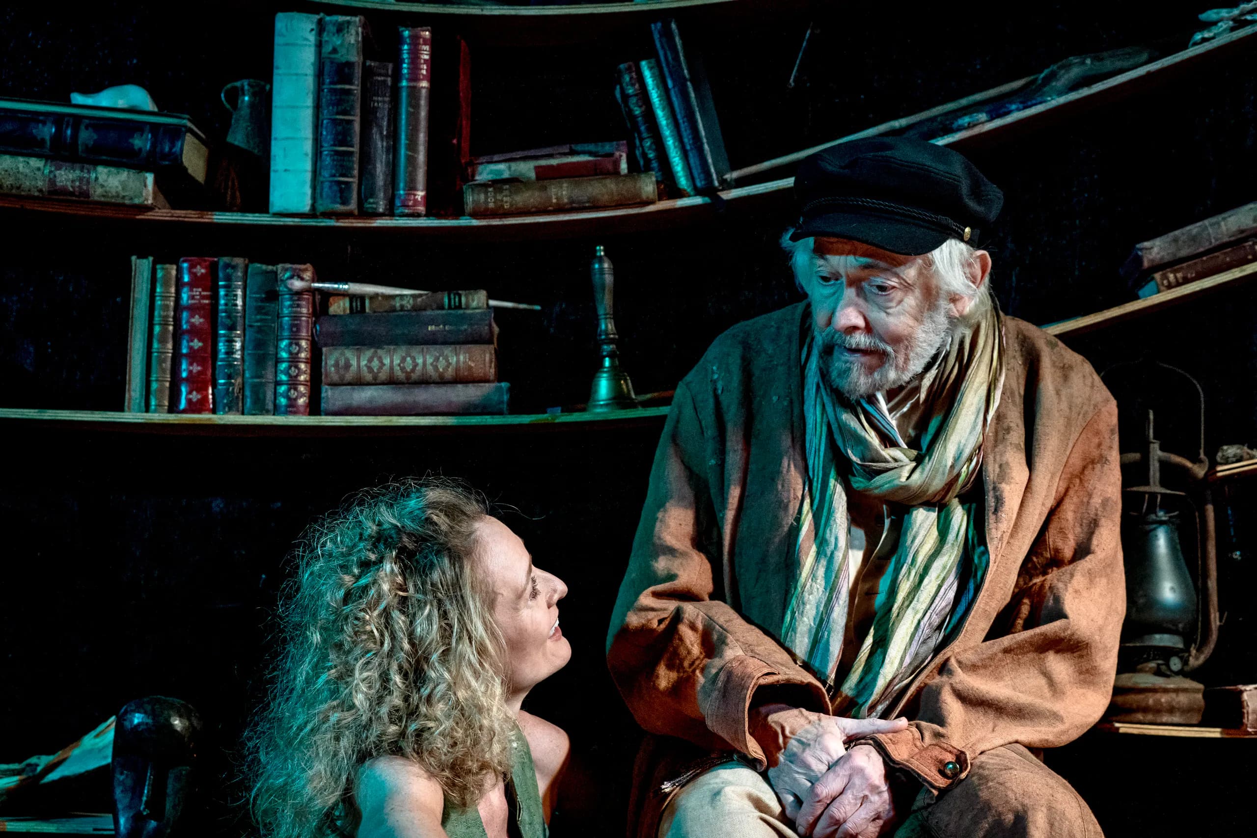 Two performers share a quiet exchange before curved bookshelves; warm, moody light in The Tempest at Jermyn Street Theatre.