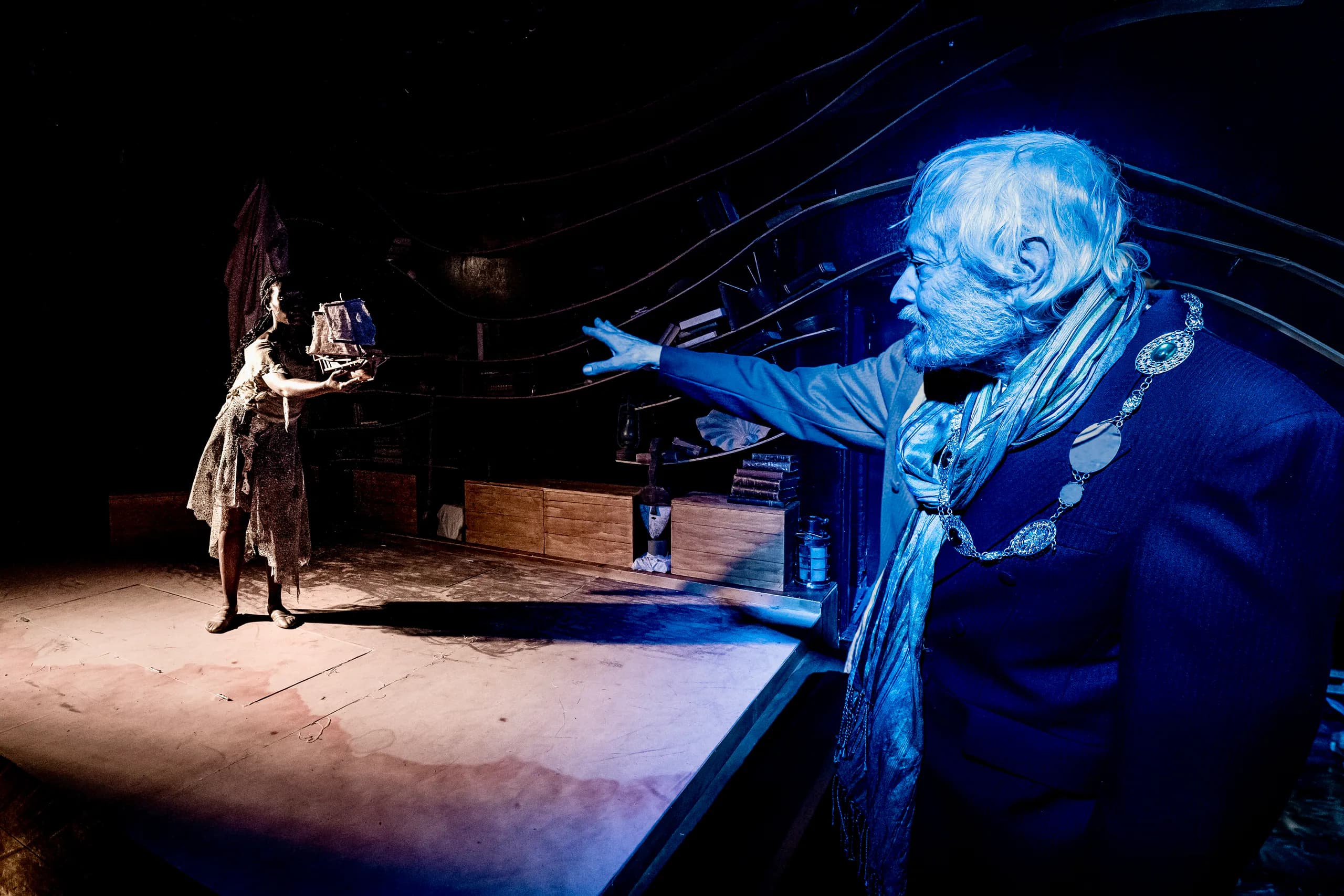 On a dim stage, an older figure in blue light reaches out as another presents a miniature ship; strong warm–cool lighting contrast.