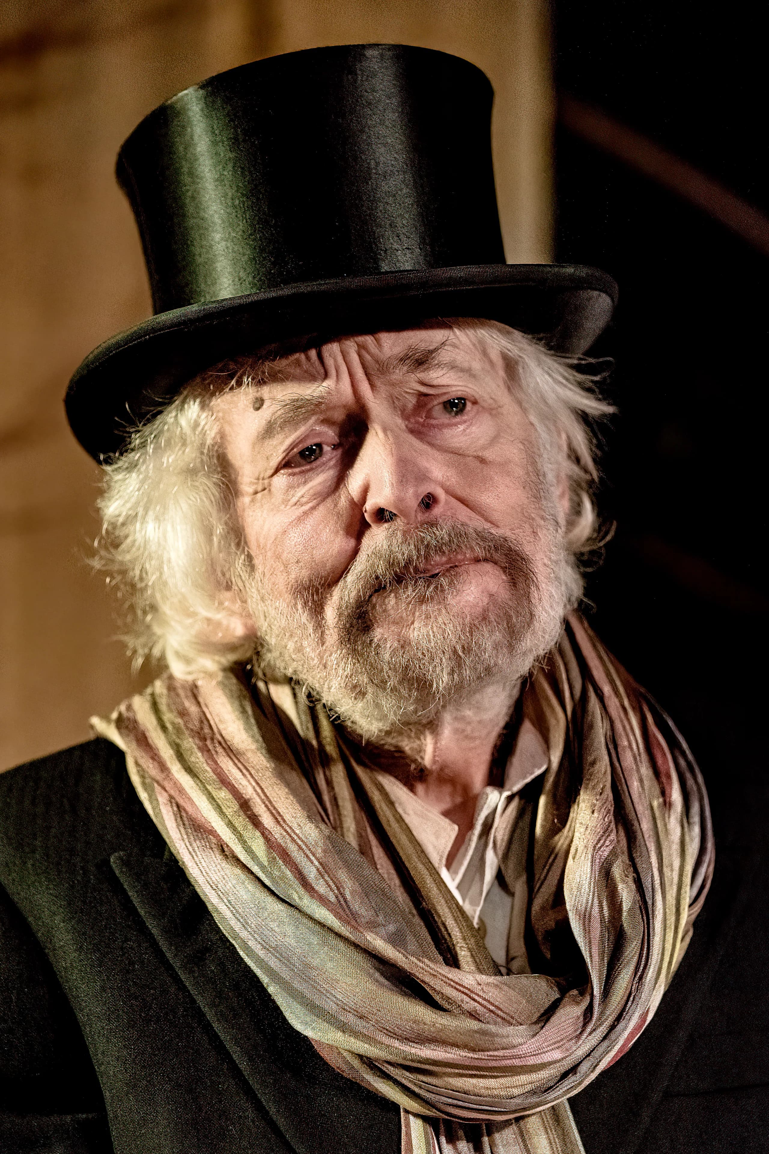 Close-up of an older performer in top hat and scarf under warm side light, reflective expression — The Tempest, Jermyn Street Theatre.
