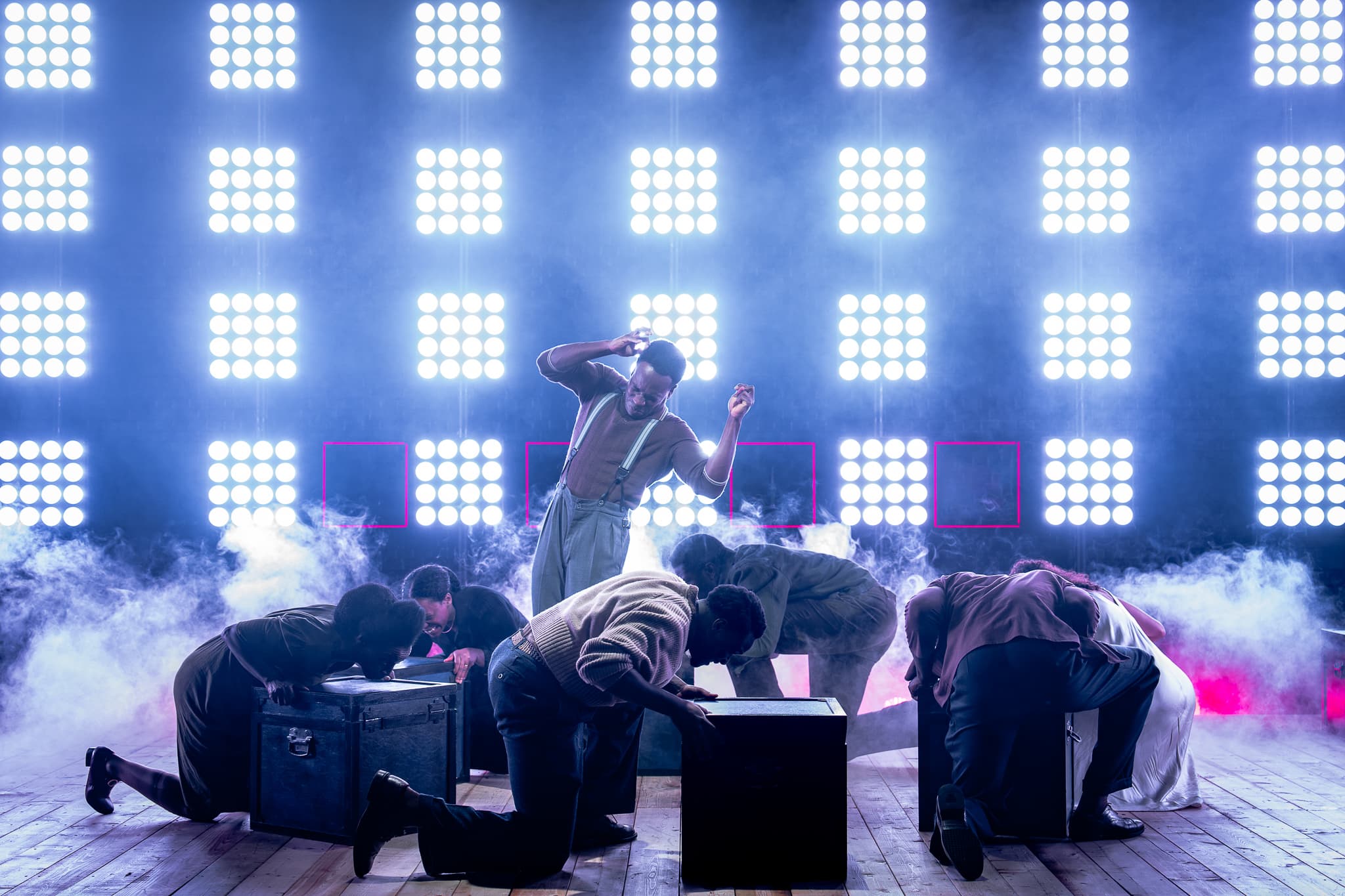 Backlit ensemble kneels over trunks as a central performer rises mid-dance amid haze and a grid of bright stage lights.