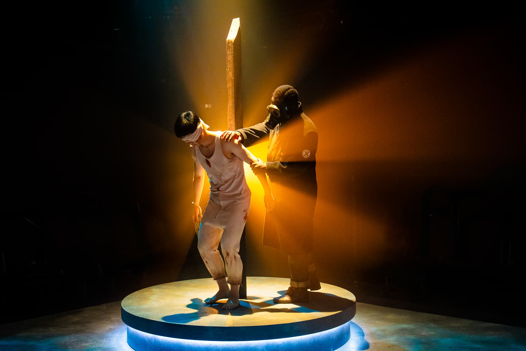 Blindfolded performer in torn whites steadied by figure in gas mask on a lit circular platform, amber backlight radiating from a post.