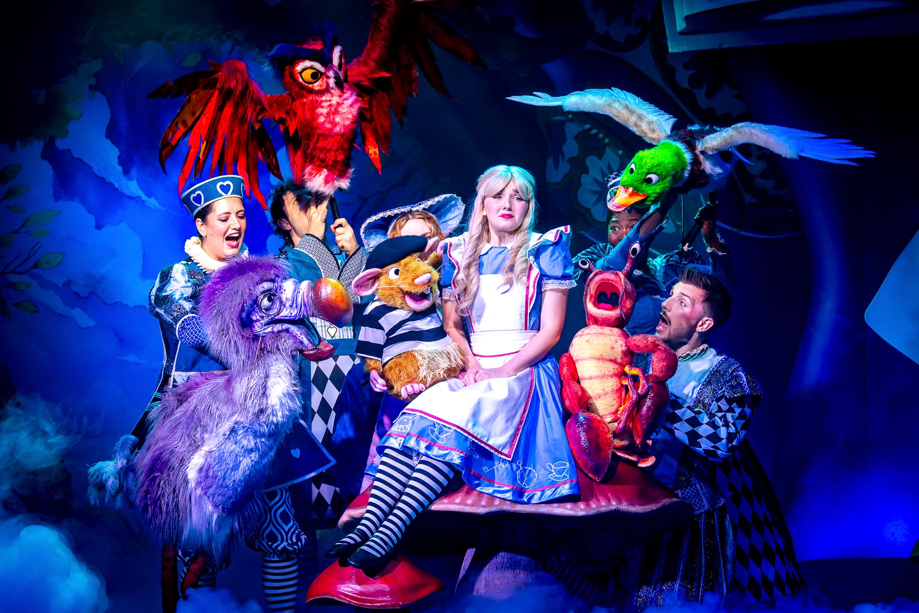 Ensemble with puppets surrounds a performer in blue dress on a giant toadstool, under vivid blue lighting and stage fog.