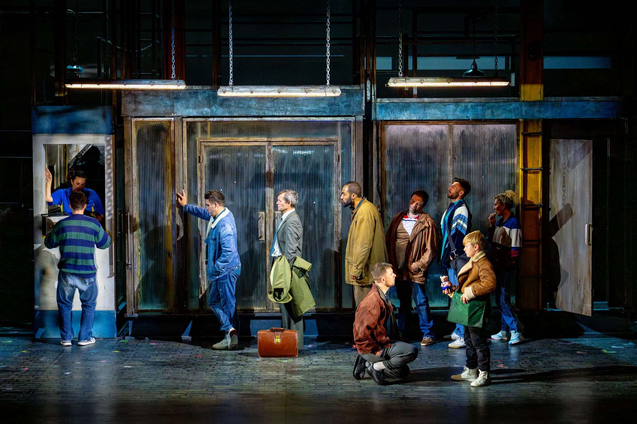 Ensemble queues outside corrugated doors under strip lights; a kneeling man speaks to a child at the front during a staged scene.