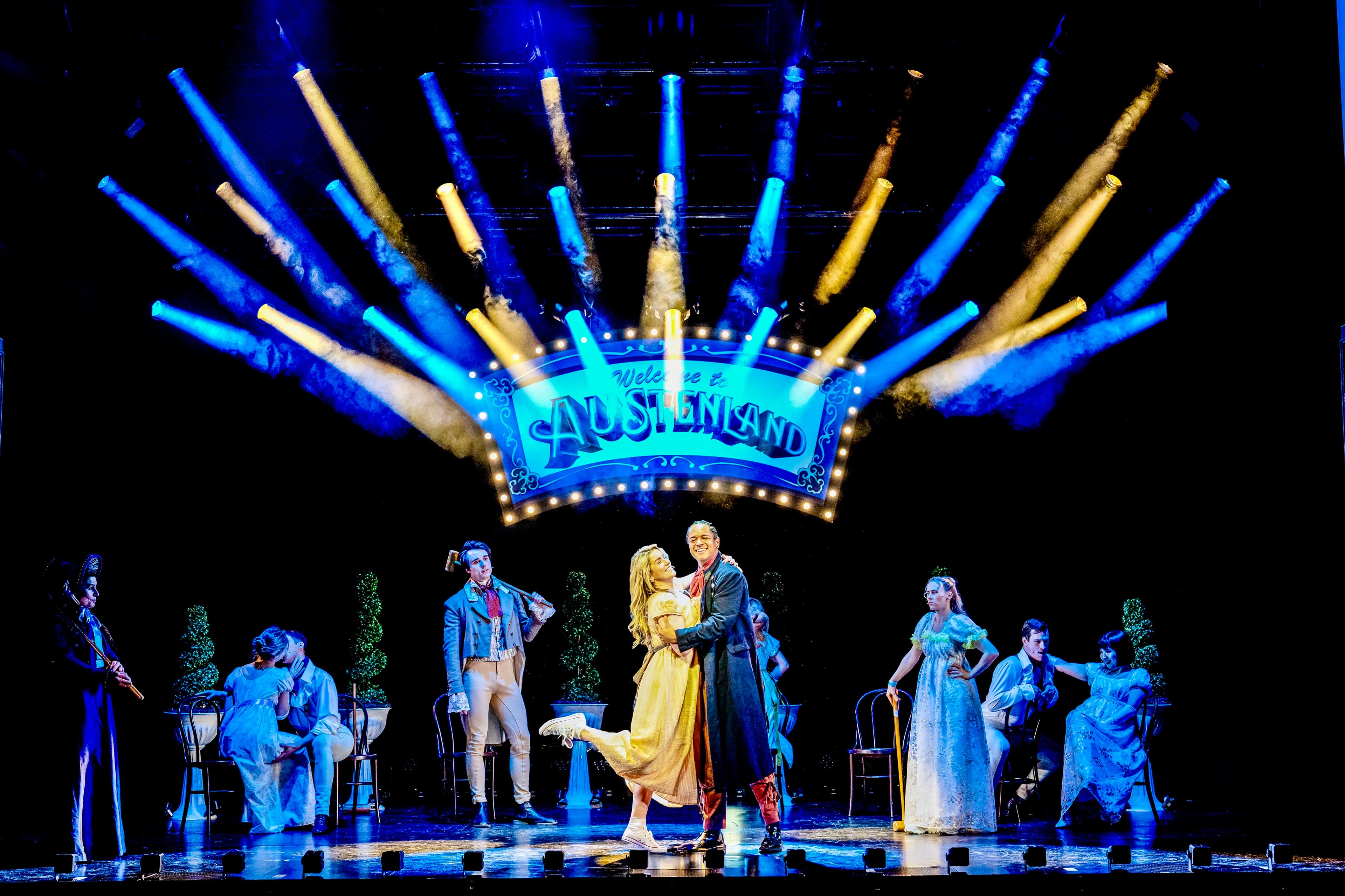 Ensemble in period costumes dancing center stage under “Welcome to Austenland” sign, with blue and gold beams and haze.