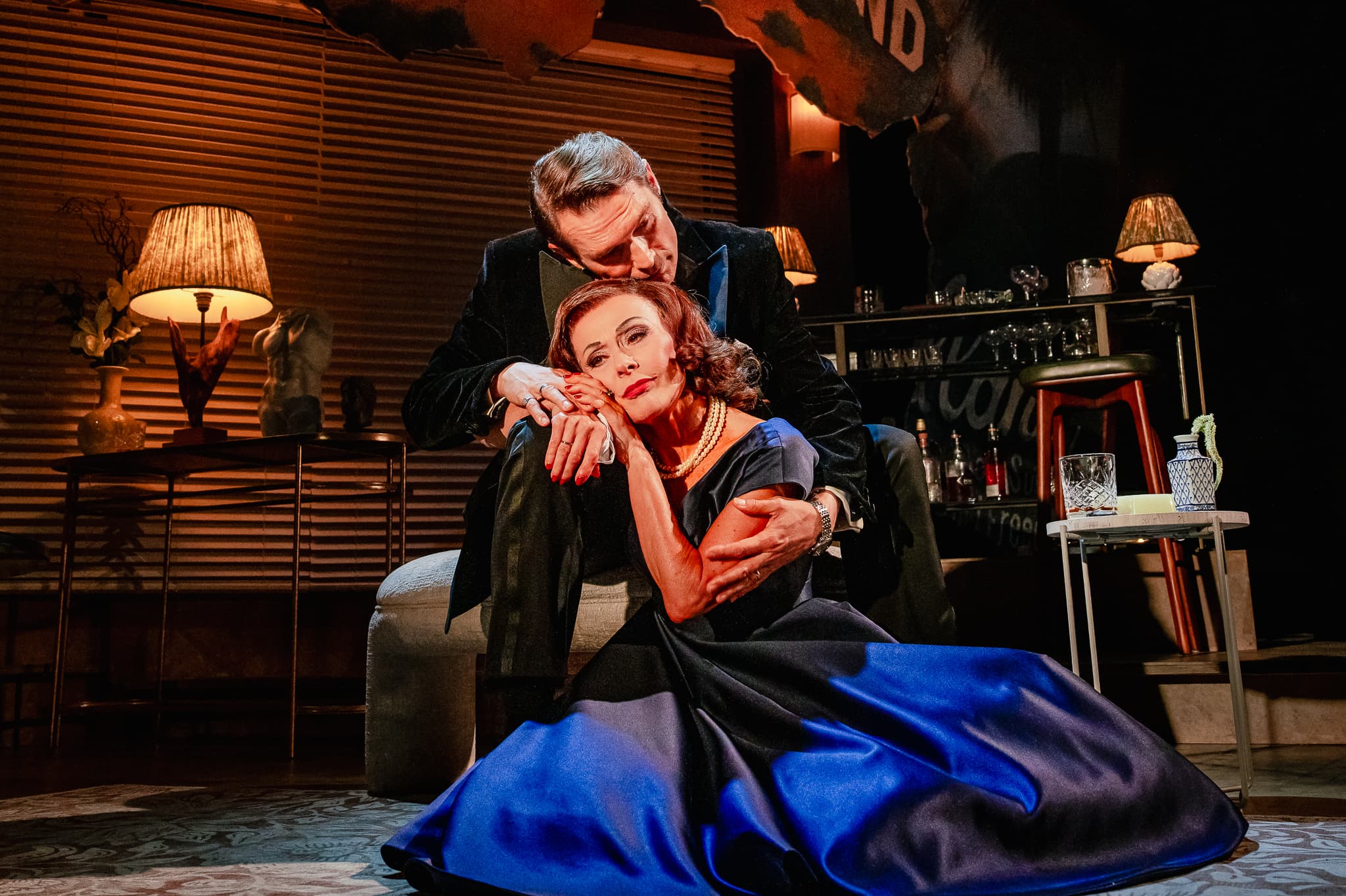 Two performers in intimate embrace on mid-century living-room set; woman in blue gown on floor, warm lamps and bar backdrop.