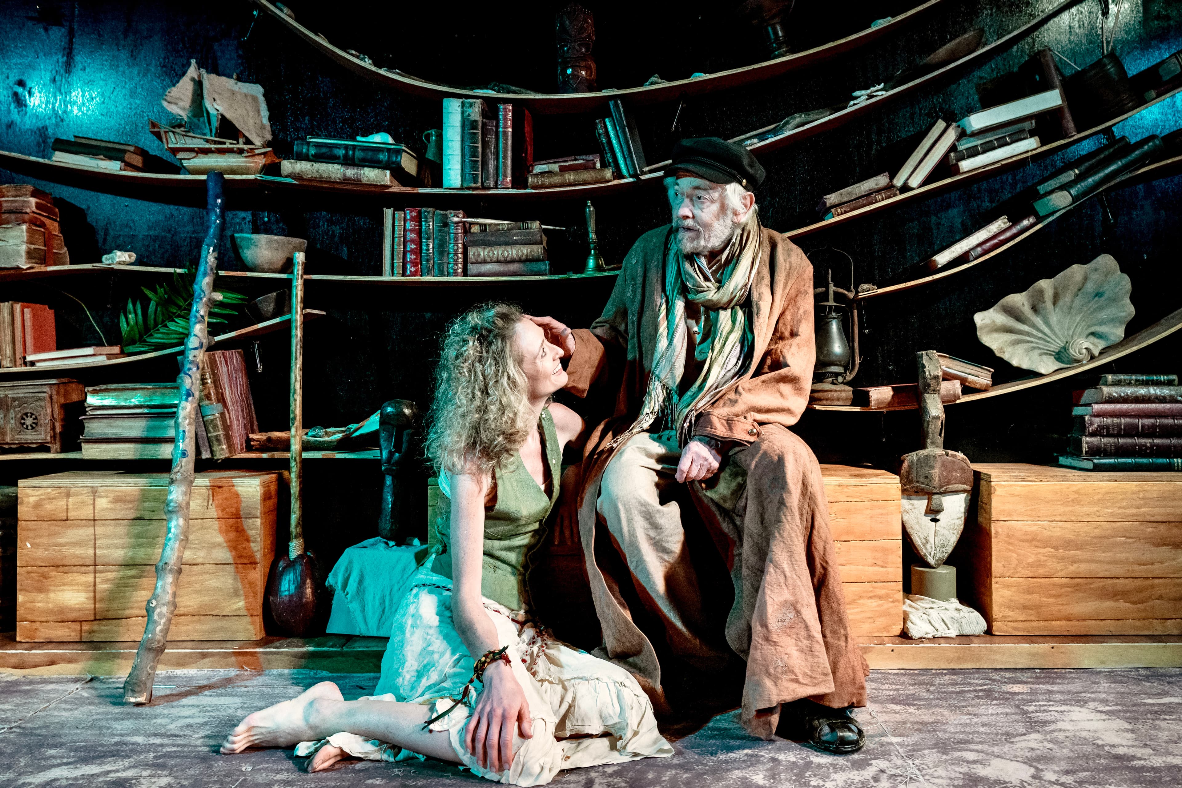 older man in coat sits as woman kneels beside him, sharing an intimate moment on stage before curved shelves of books, shells, and nautical props