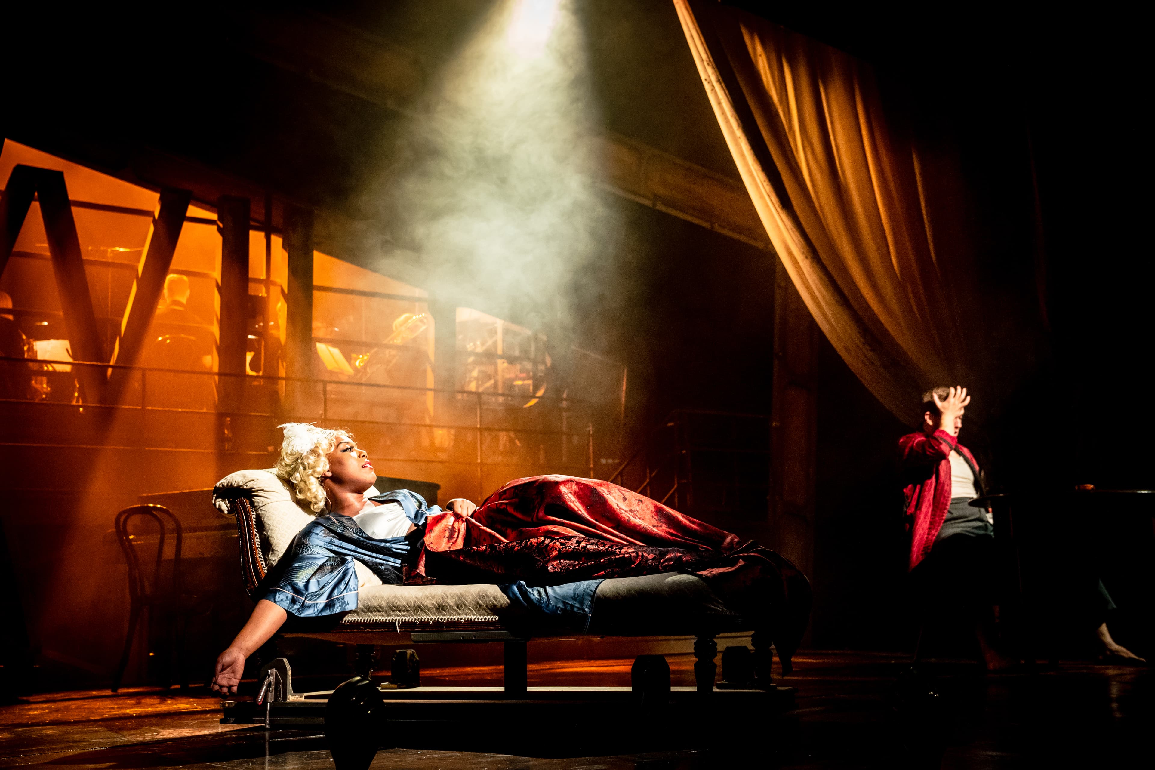 Performer in powdered wig reclines on chaise under spotlight and haze; second figure in red robe in shadow; band backlit behind railing.