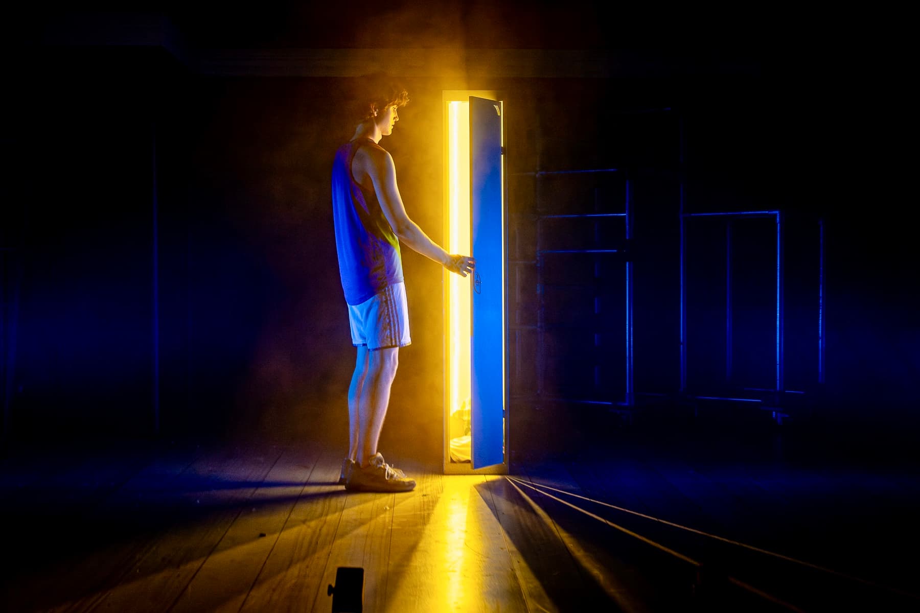 Performer in sportswear opens a glowing door on a dark stage, blue and amber haze cutting across the floor.