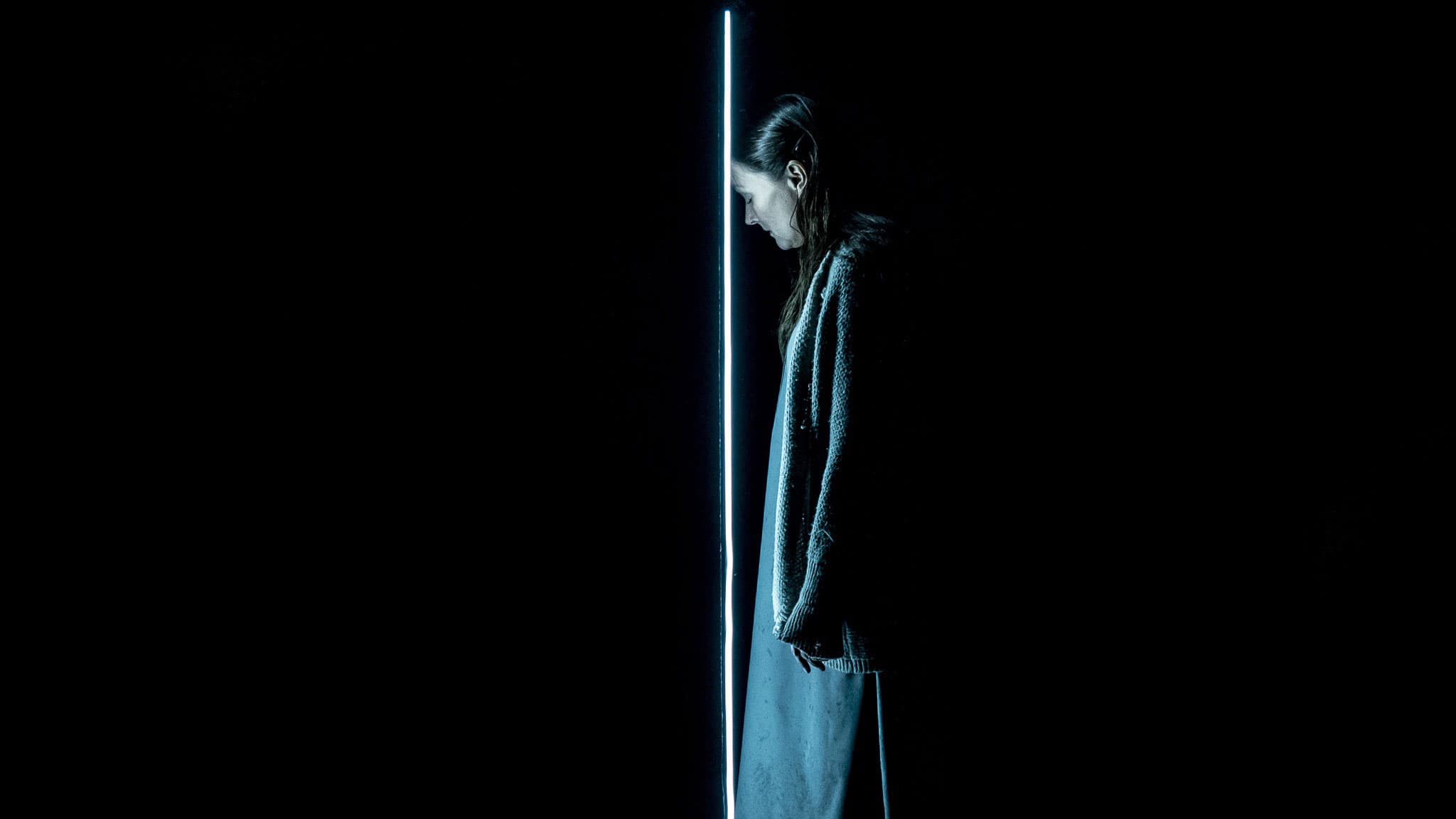 performer in profile beside a vertical light strip, isolated in blue light against black stage; minimalist staging and somber mood