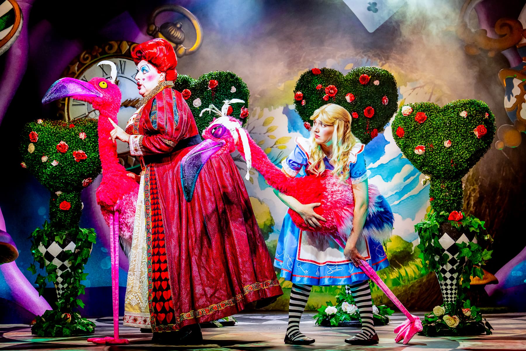 Two actors play croquet with pink flamingo props amid heart-shaped topiaries, vivid costumes, and saturated stage lighting.