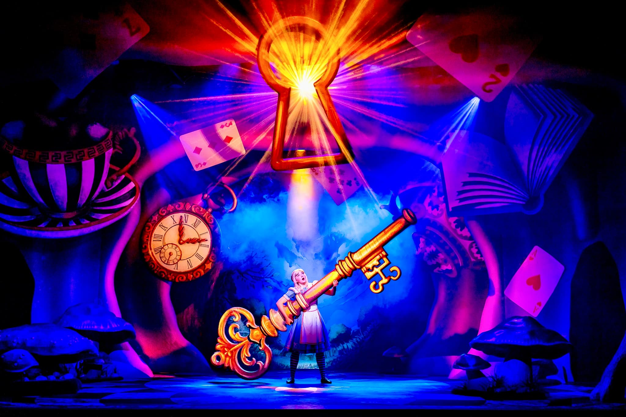 Performer in blue dress lifts an oversized golden key under a glowing keyhole amid cards, clock, mushrooms and book; vivid blue-magenta lighting.