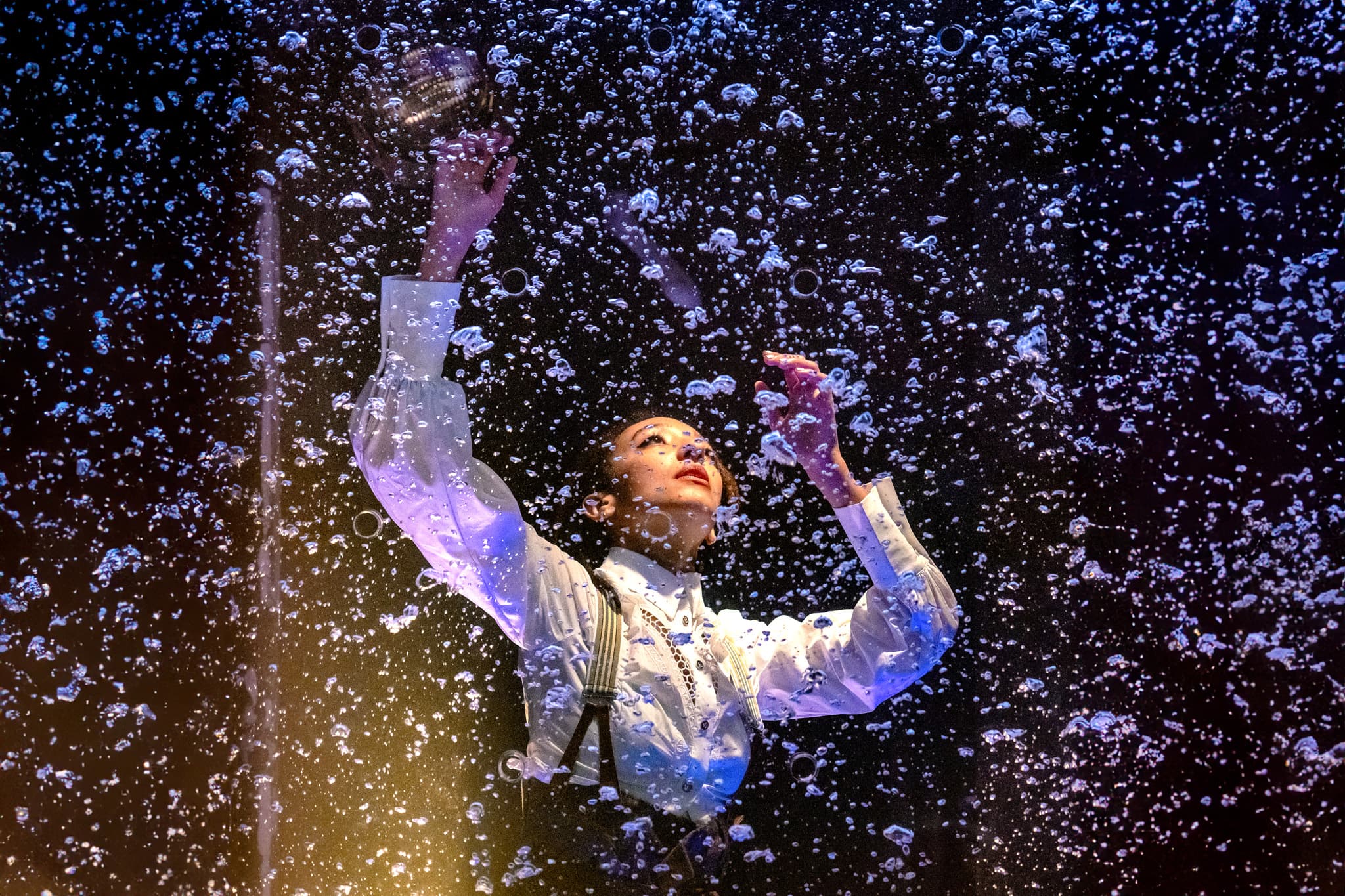 performer in white shirt and suspenders reaching toward a rain-splattered screen under blue stage lighting