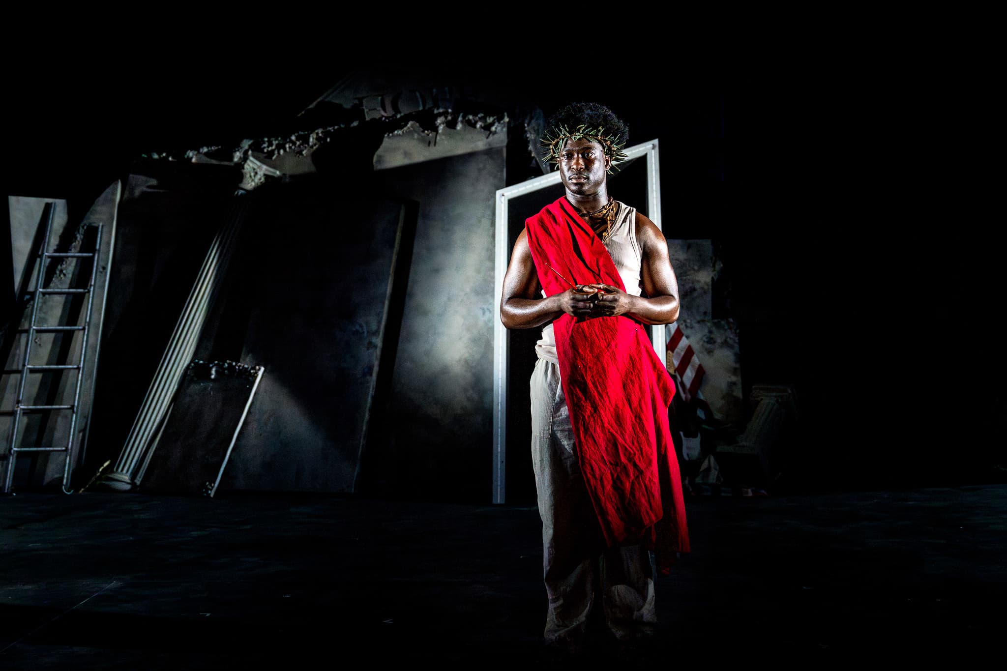 Performer center stage in red drape and thorn crown, spotlighted against a dark, ruined set with fallen columns and a ladder.