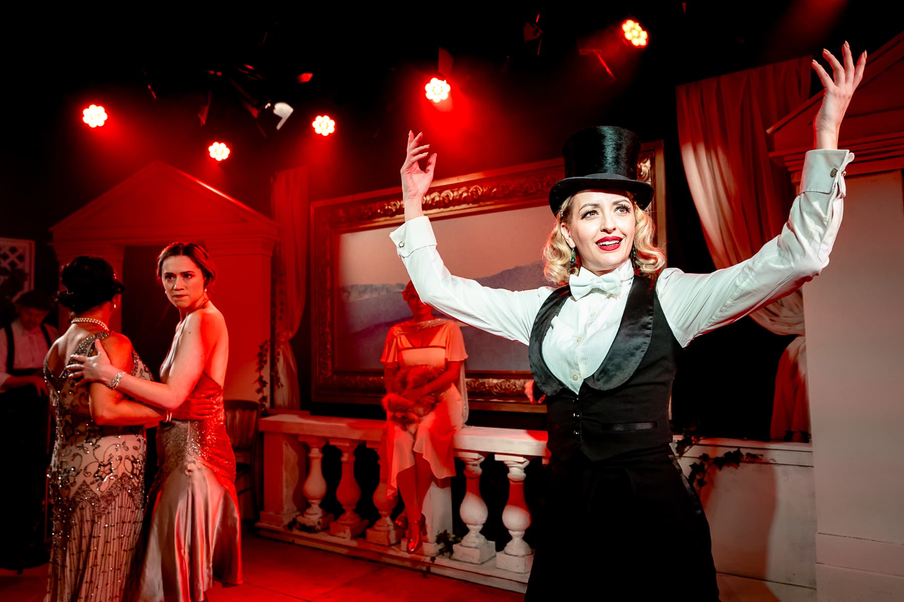 Top-hatted performer raises arms in foreground; two dancers hold at left under red stage lights against an art deco set.