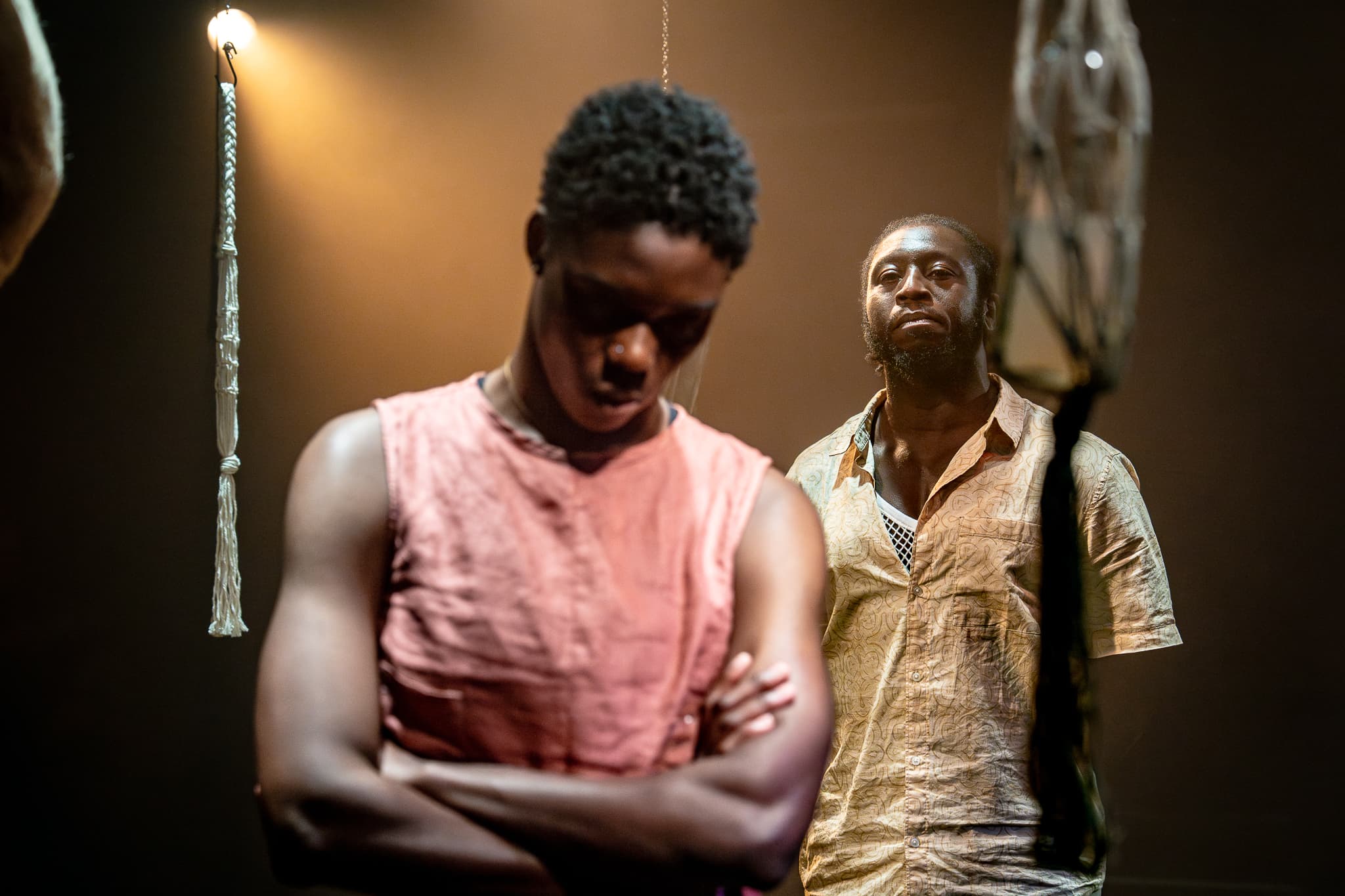 Two performers on stage; foreground figure with arms crossed and head bowed, background figure watching; warm spotlight and hanging bulb.
