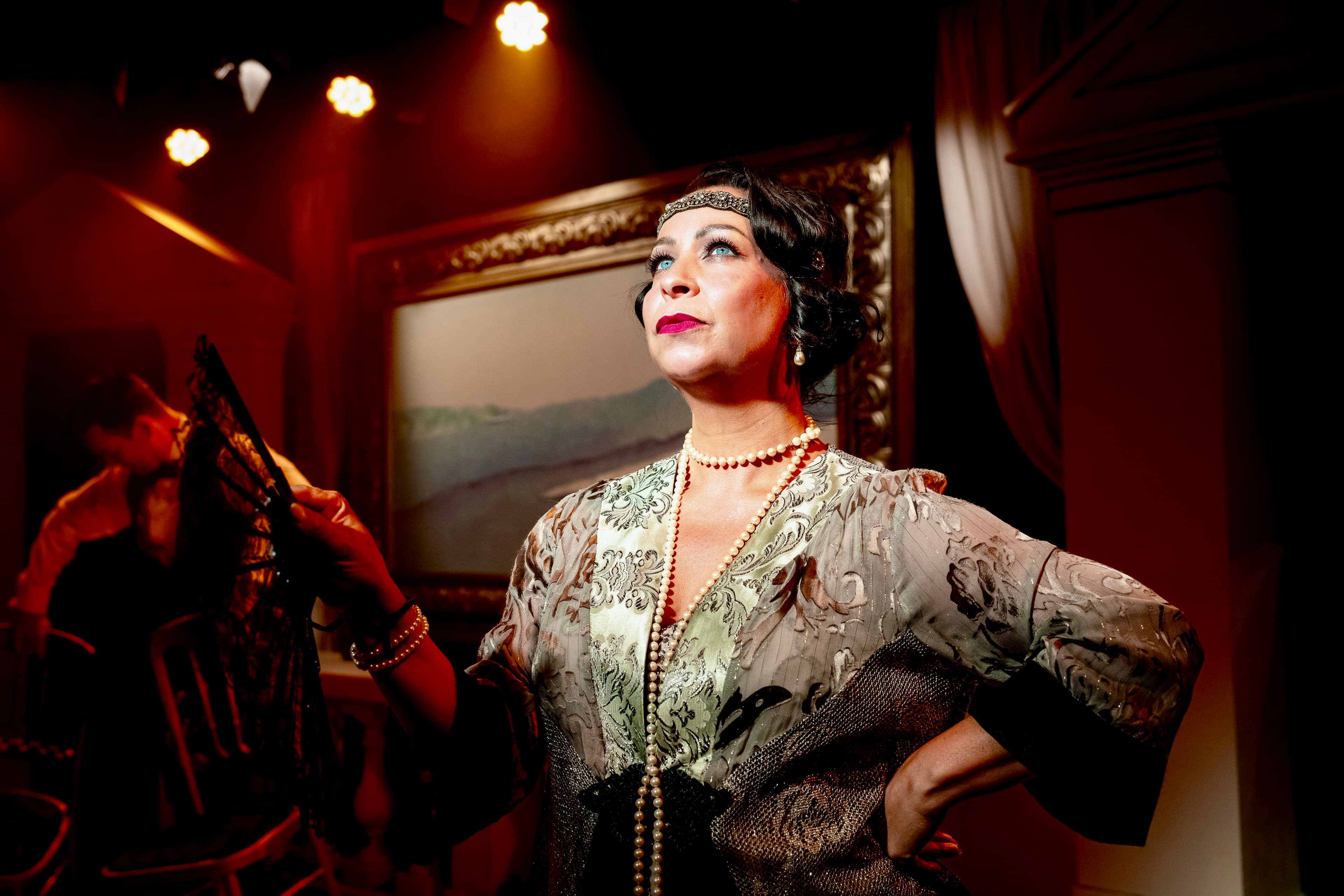performer in vintage-style beaded dress and pearls holds a lace fan under warm stage lights; framed painting and seated figure behind