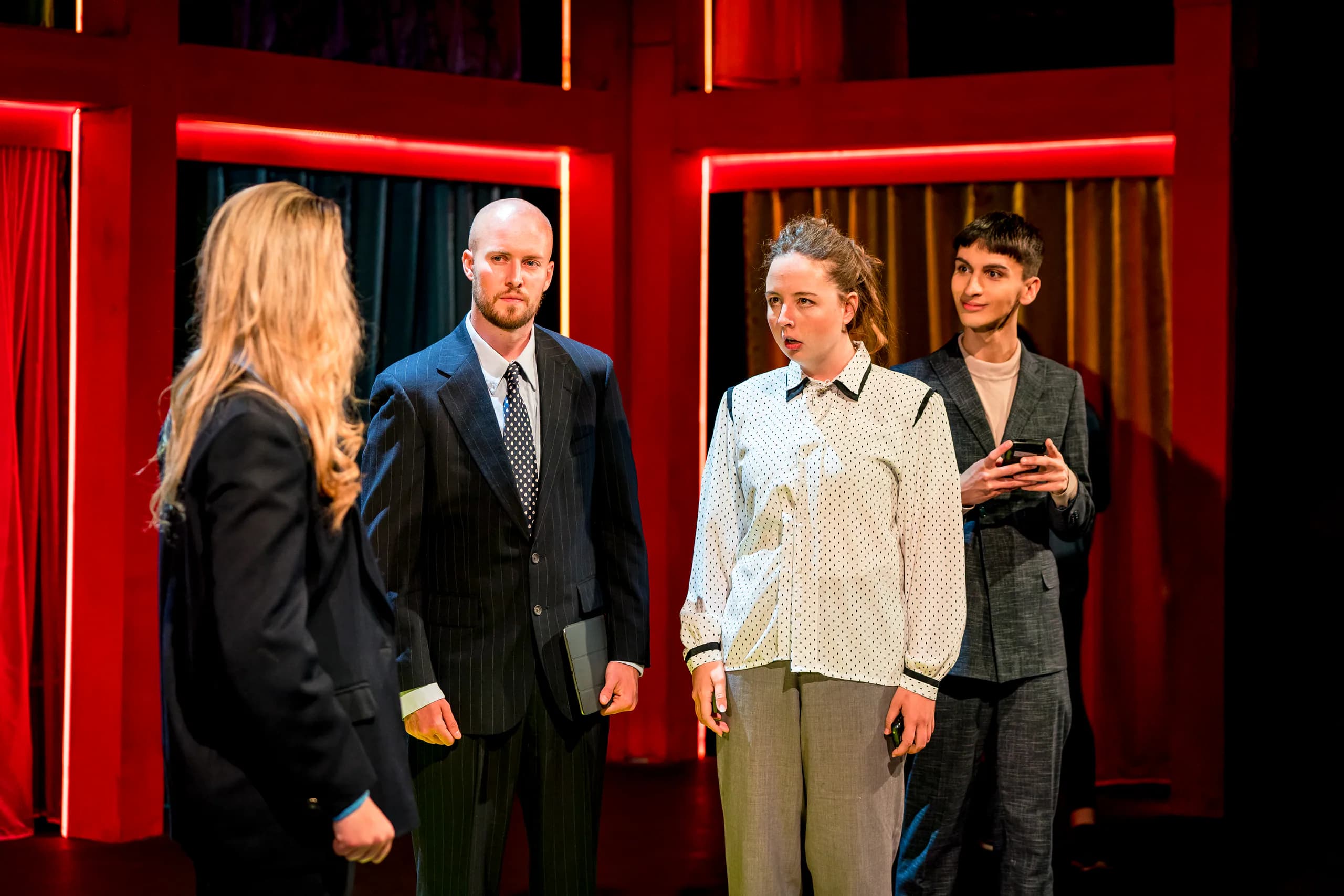 Four actors confer on a red-framed set, warm lighting highlighting a tense exchange in the GSA production of 13.