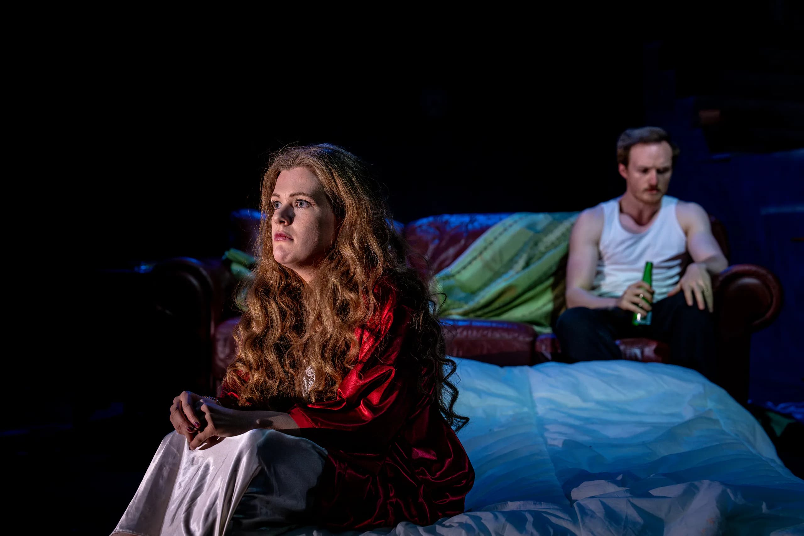 Woman in red robe sits pensively on a bed; man in vest with beer on sofa behind under moody blue light.