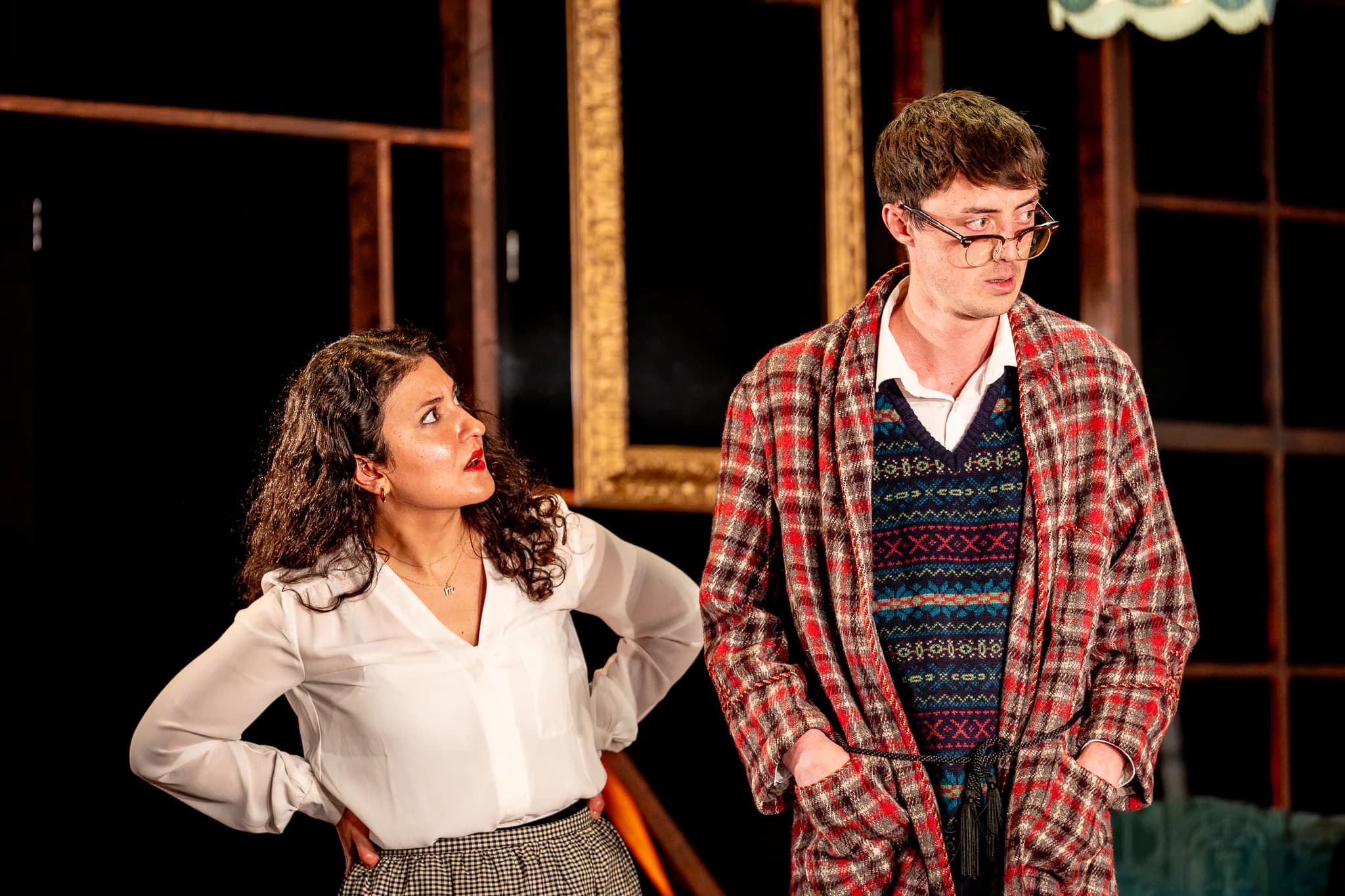 Man in glasses, robe and knit vest stands beside woman with hands on hips on a warmly lit domestic set.