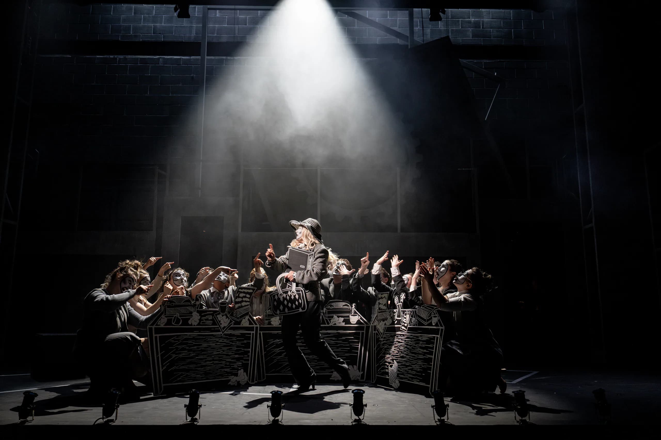 Central performer in spotlight as ensemble crowds around office desks; dramatic, smoky lighting in 9 to 5 at Mountview.