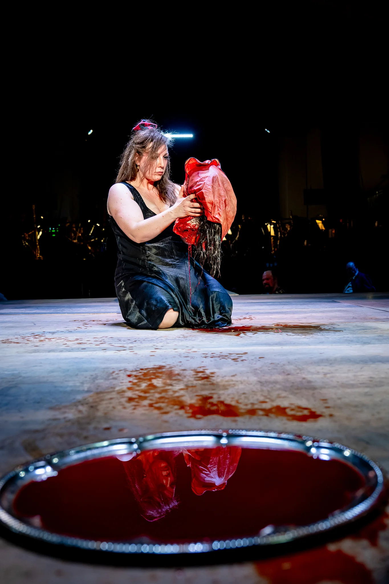 Salome kneels on a blood-spattered stage holding the veiled head, her reflection caught in the pool of blood on a silver platter.